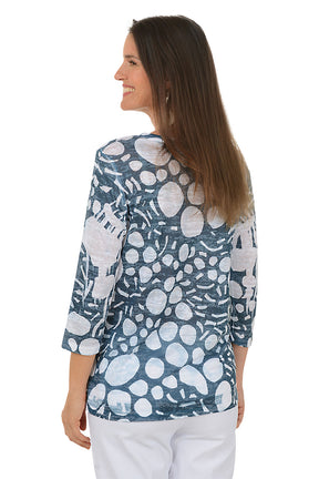 Woman wearing a blue floral patterned top with white pants on a white background. Back shot.