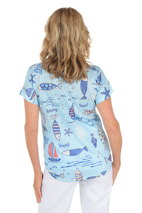 A woman wearing a blue short sleeve v-neck shirt with a fish and sailboat design. Back shot.