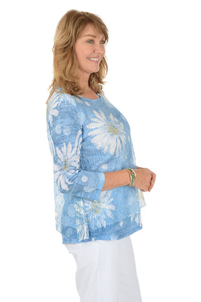 A woman wearing  a blue 3/4 sleeve shirt with a daisy pattern detail. Side shot.