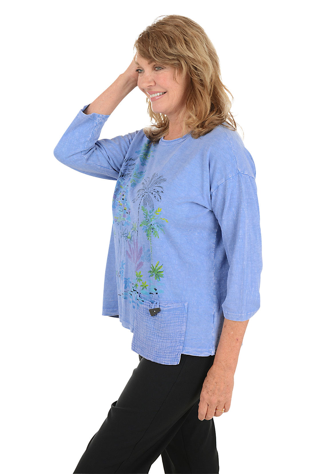 A woman wearing a 3/4 sleeve blue top with a palm tree pattern detail. Side shot.