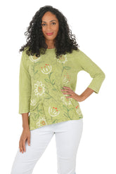 Woman wearing a green floral blouse with white pants on a white background.