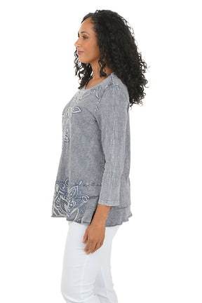 Woman wearing a gray floral-patterned top and white pants on a white background. Side shot.