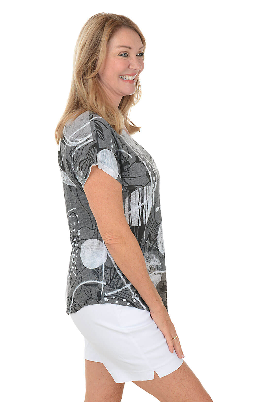 A woman wears a printed grey and white t-shirt and white shorts against a white background. Side shot.