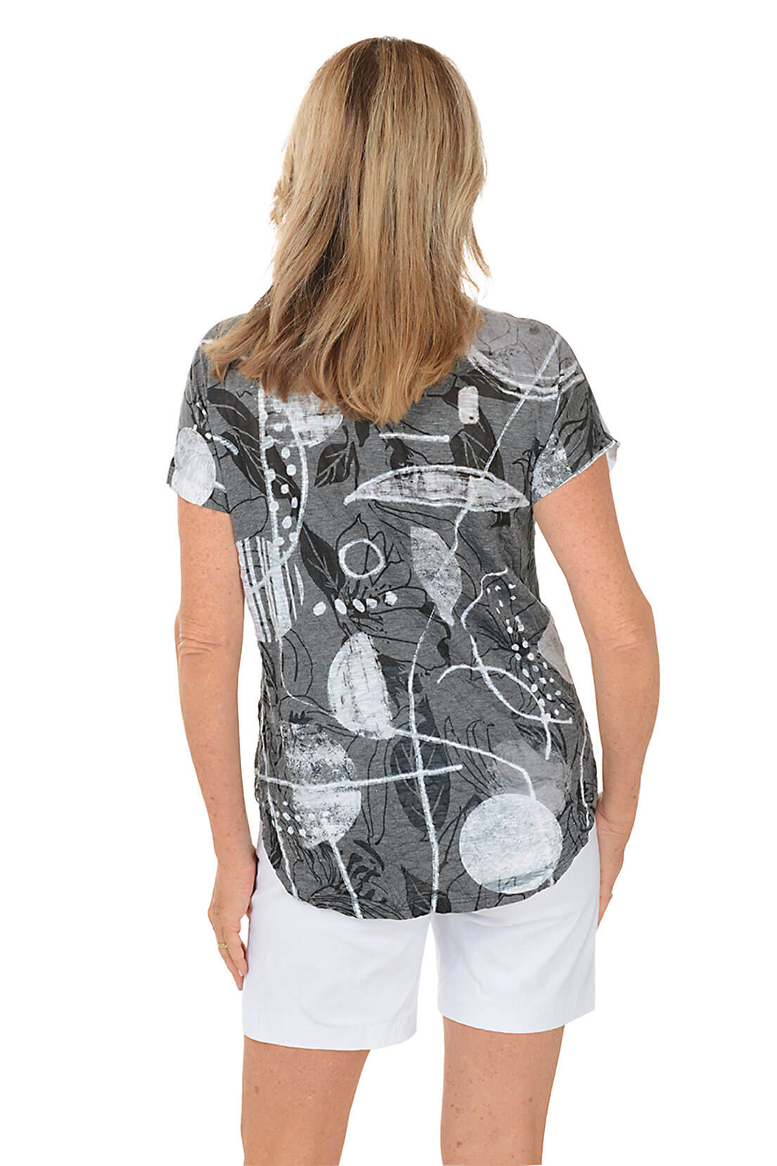 A woman wears a printed grey and white t-shirt and white shorts against a white background. Back shot.