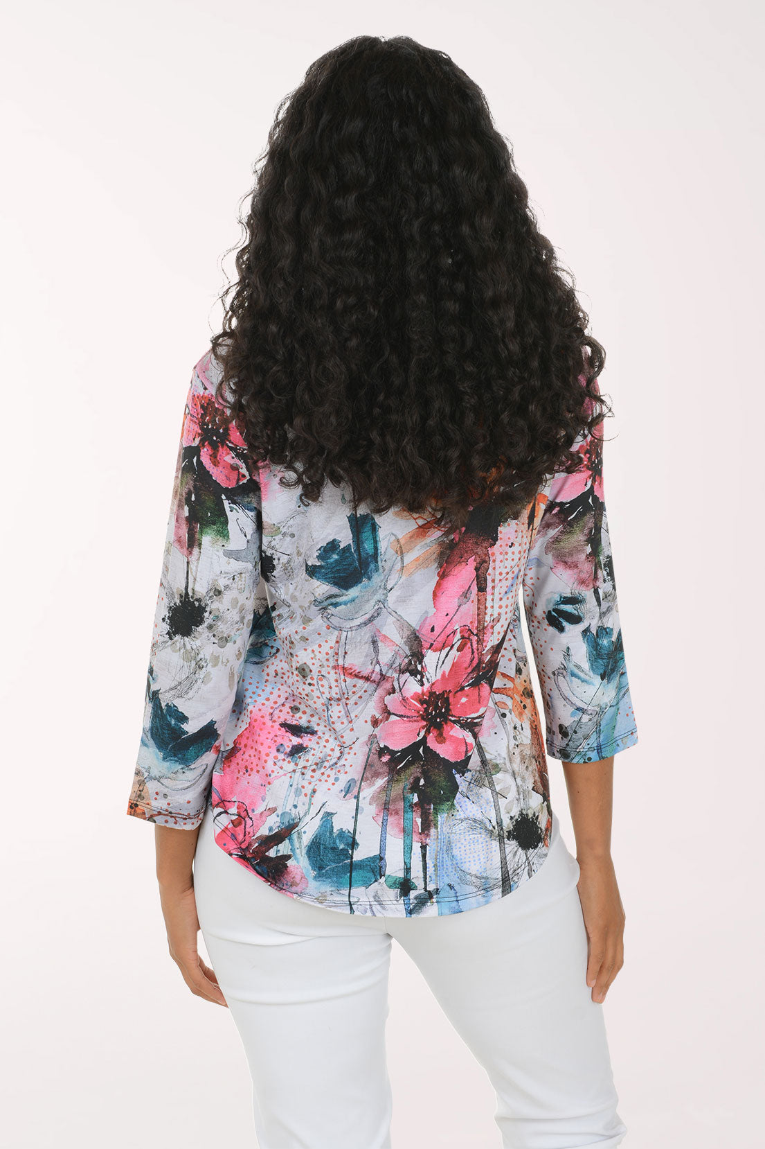 A woman with long curly black hair wearing a colorful abstract floral print top and white pants, standing against a plain white studio background.