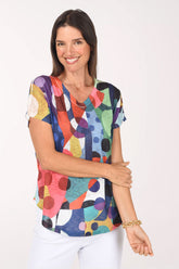 Model wearing the Jess and Jane Dot Day Slub Knit Short Sleeve Top, bold dots and color blocks in red, coral, purple, yellow, teal and navy, over white pants, front view