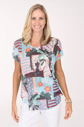 Model wearing the Jess and Jane Sunny Day Slub Knit Short Sleeve Top, a pastel patchwork of palms and beach scenes, over white pants, front view
