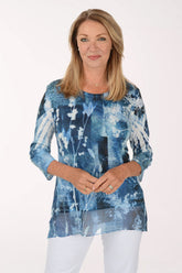 A woman modeling a blue and white abstract patterned long-sleeve tunic top with white pants, photographed against a plain white studio background.