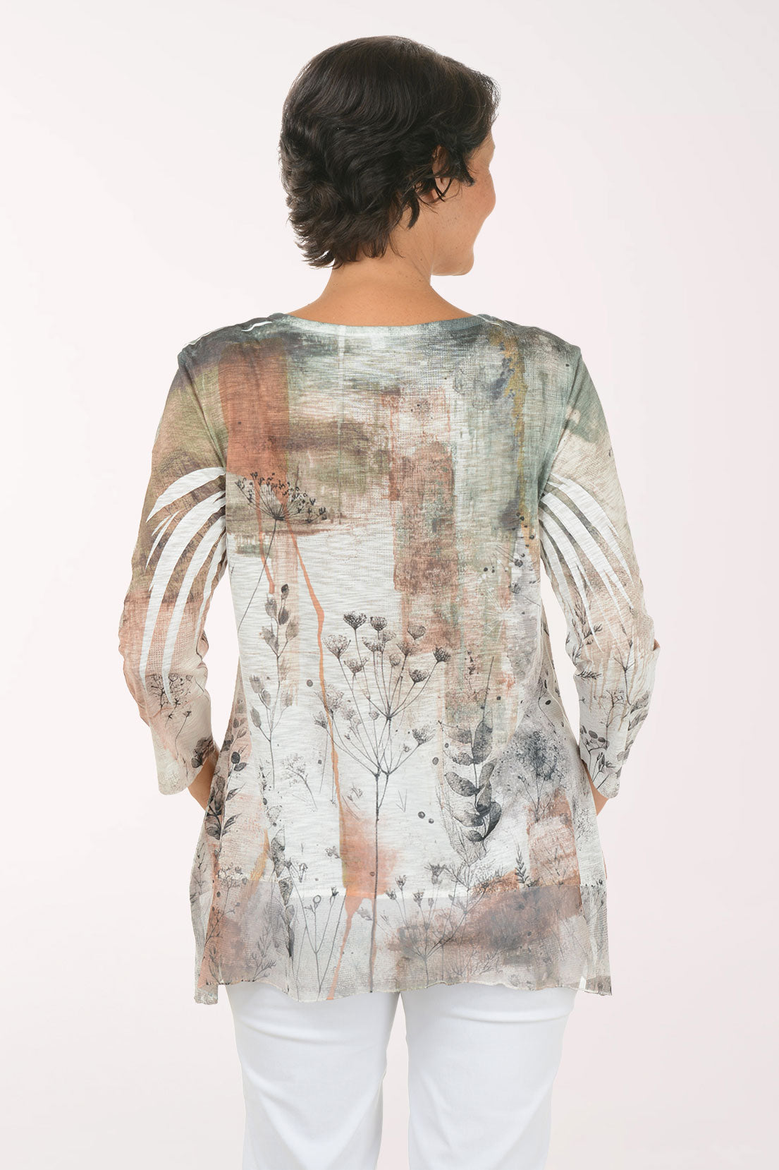 A woman modeling a three-quarter sleeve tunic top with an abstract botanical print in muted tones of beige, gray, brown, and green, paired with white pants, against a plain white studio background.