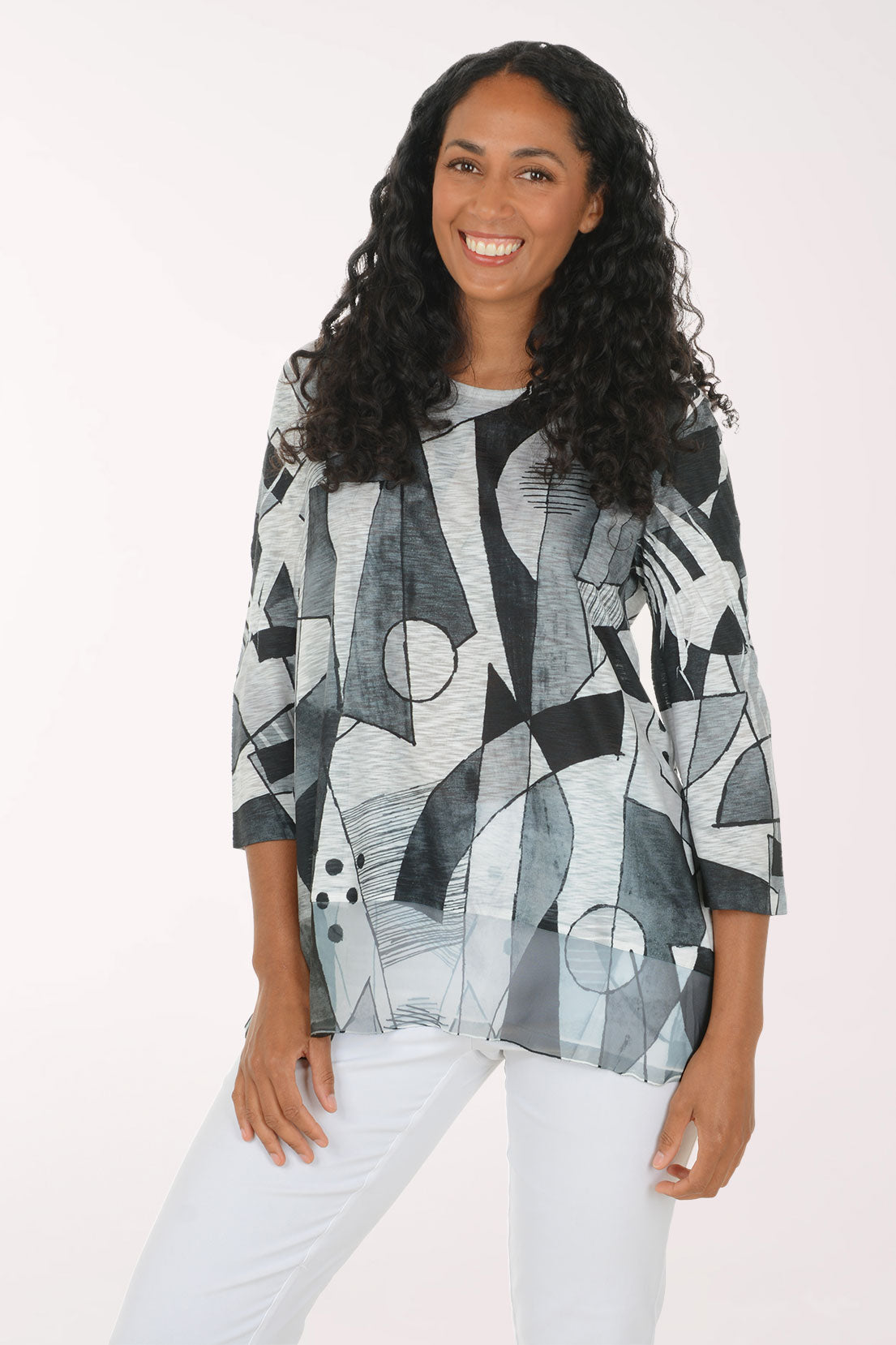 A woman with long curly dark hair wearing a loose-fitting, three-quarter sleeve top with an abstract geometric print in black, gray, and white, paired with white pants, against a plain light background.
