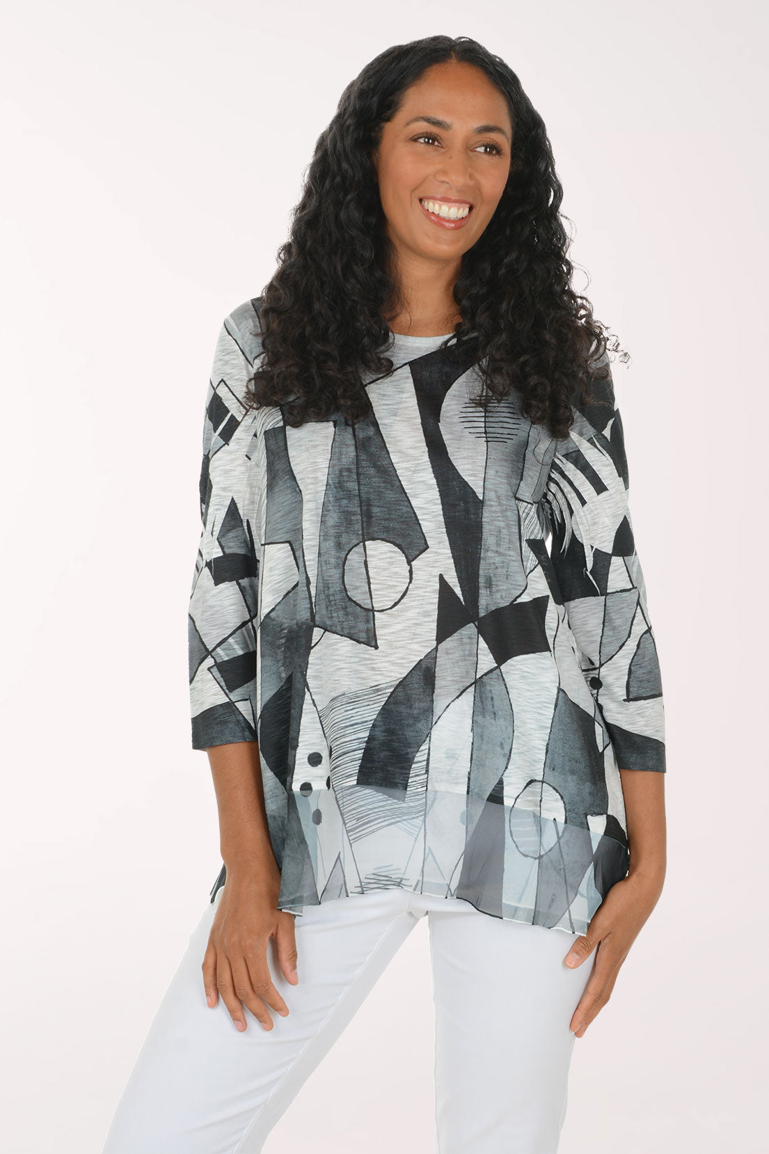 A woman with long curly dark hair wearing a loose-fitting, three-quarter sleeve top with an abstract geometric print in black, gray, and white, paired with white pants, against a plain light background.