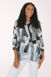 A woman with long curly dark hair wearing a loose-fitting, three-quarter sleeve top with an abstract geometric print in black, gray, and white, paired with white pants, against a plain light background.