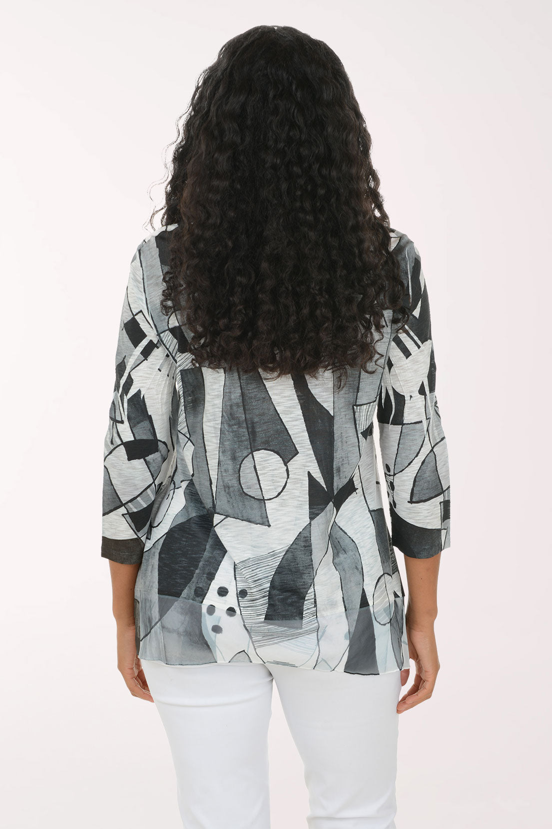 A woman with long curly dark hair wearing a loose-fitting, three-quarter sleeve top with an abstract geometric print in black, gray, and white, paired with white pants, against a plain light background.
