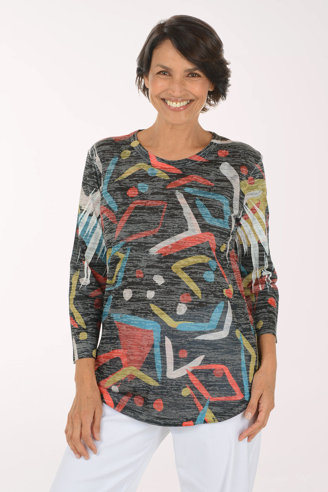 A smiling woman wearing a long-sleeve abstract print top with white pants, photographed against a plain light background.