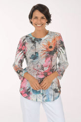 A smiling woman wearing a long-sleeve abstract floral print top with pink, orange, blue, and gray tones, paired with white pants, against a plain white studio background.