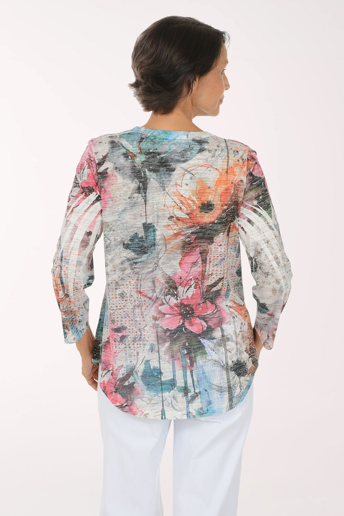 A smiling woman wearing a long-sleeve abstract floral print top with pink, orange, blue, and gray tones, paired with white pants, against a plain white studio background.