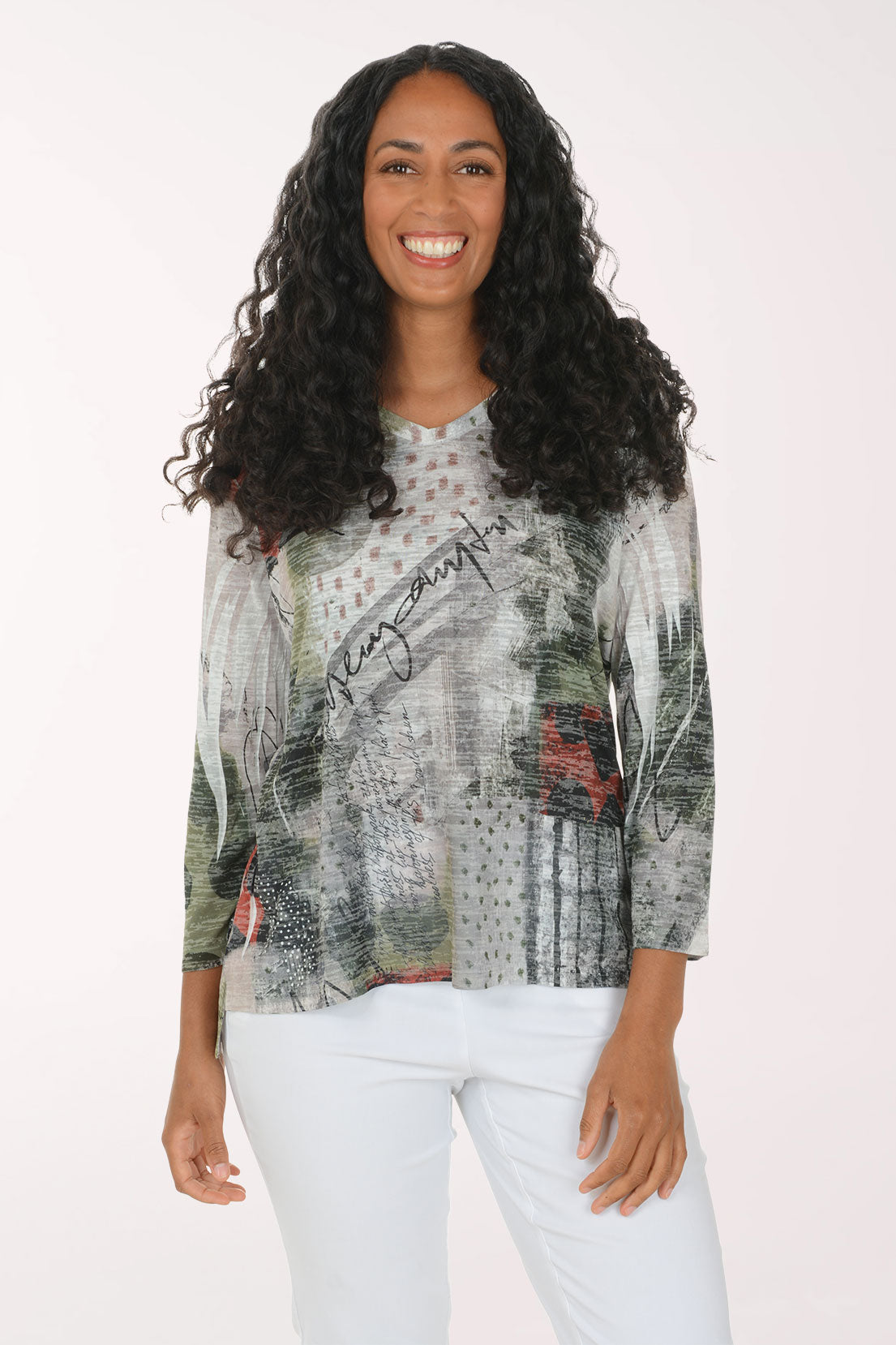 A woman with long, curly black hair is smiling and wearing a three-quarter sleeve top with an abstract, painterly print in gray, green, black, and red, paired with white pants, against a plain white studio background.
