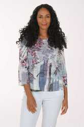 A woman with long, curly black hair wearing a three-quarter sleeve blouse with a blue, purple, and pink abstract botanical print, paired with white pants, standing against a plain white studio background.