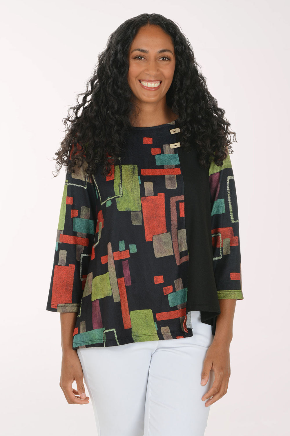 A smiling woman with long curly black hair wearing a black three-quarter sleeve top with a colorful geometric block pattern in red, green, and beige, paired with white pants, against a plain white studio background.