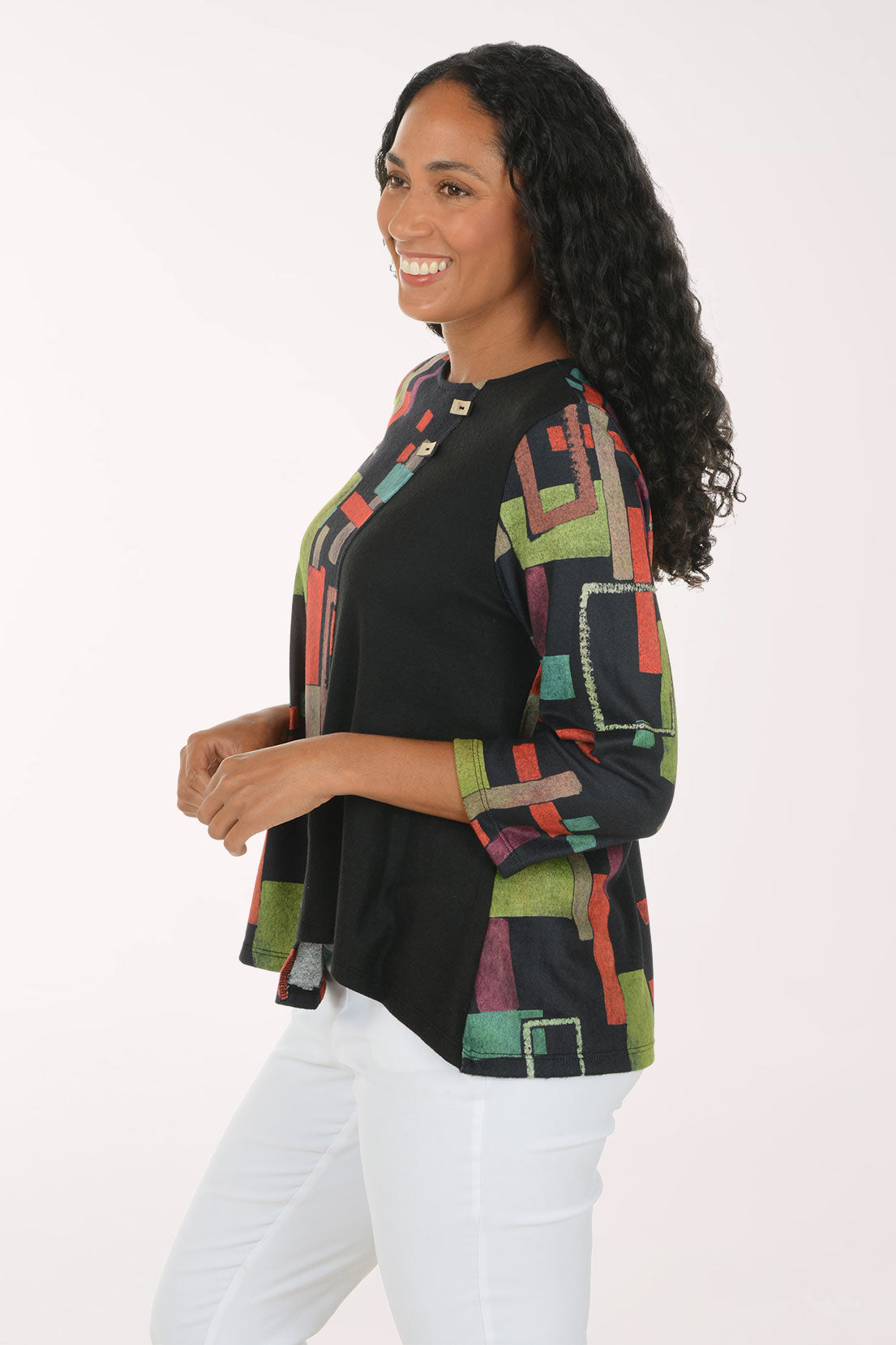 A smiling woman with long curly black hair wearing a black three-quarter sleeve top with a colorful geometric block pattern in red, green, and beige, paired with white pants, against a plain white studio background.