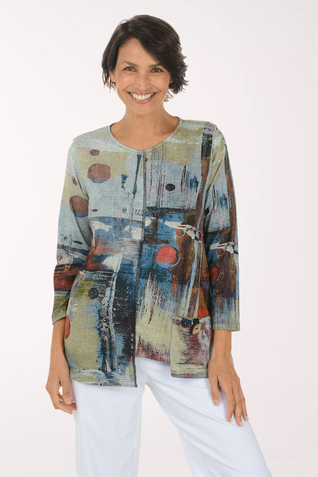 A woman modeling a long-sleeve abstract print top with blue, beige, brown, and red tones, paired with white pants, against a plain light background.