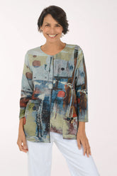 A woman modeling a long-sleeve abstract print top with blue, beige, brown, and red tones, paired with white pants, against a plain light background.