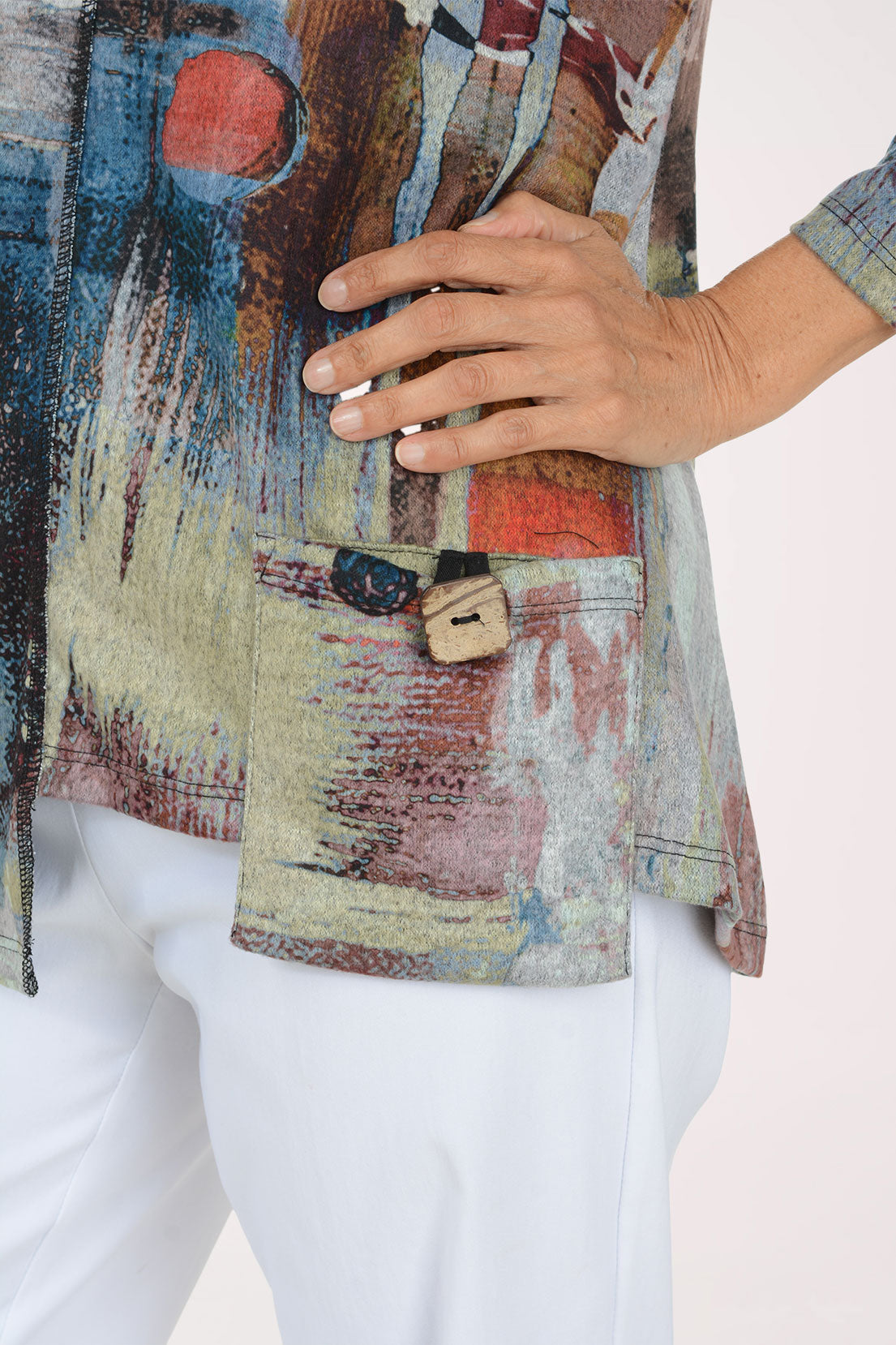 A woman modeling a long-sleeve abstract print top with blue, beige, brown, and red tones, paired with white pants, against a plain light background.