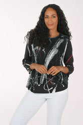 A woman modeling a black long-sleeve top with a white and gray abstract brushstroke pattern and thin red accent lines, paired with white pants, against a plain white studio background.