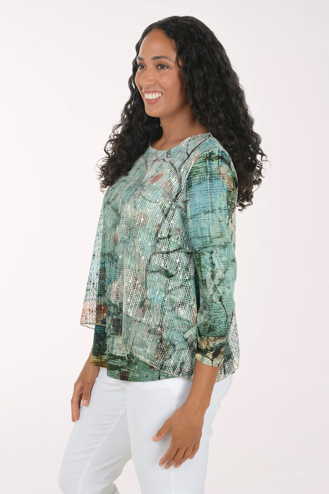 A woman with long, curly black hair wearing a green and blue abstract-patterned blouse and white pants, standing against a plain white studio background.
