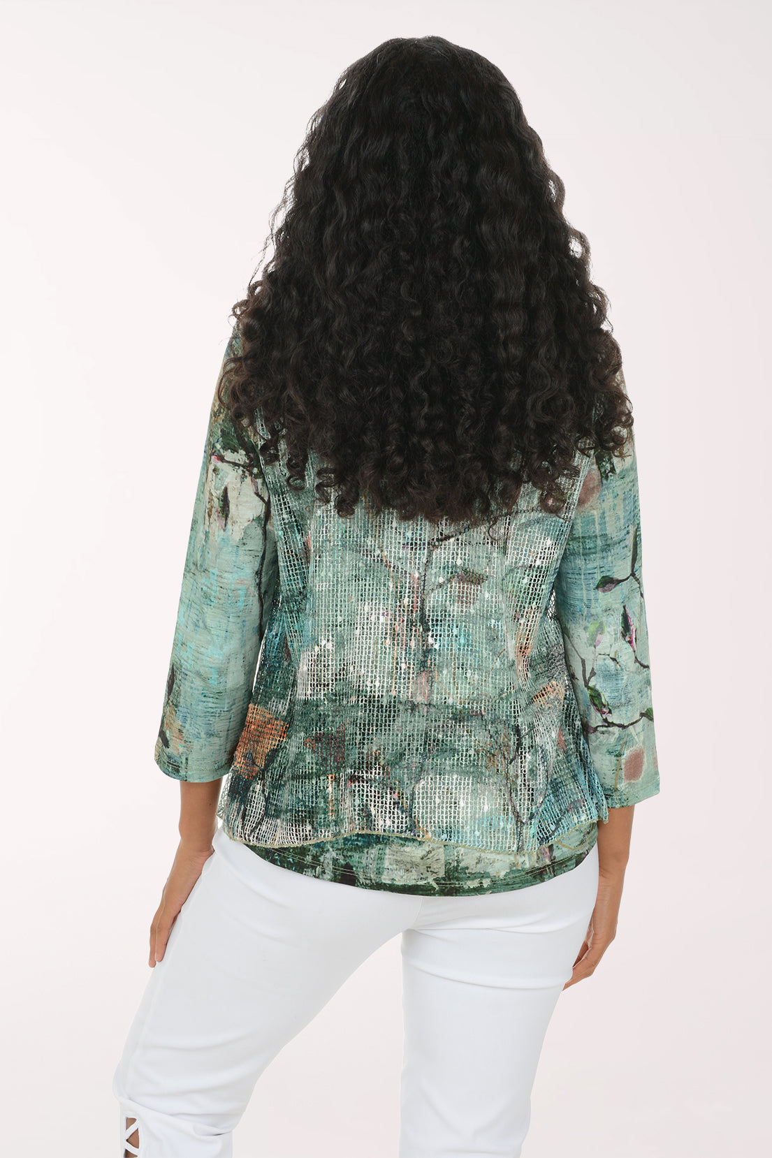 A woman with long, curly black hair wearing a green and blue abstract-patterned blouse and white pants, standing against a plain white studio background.
