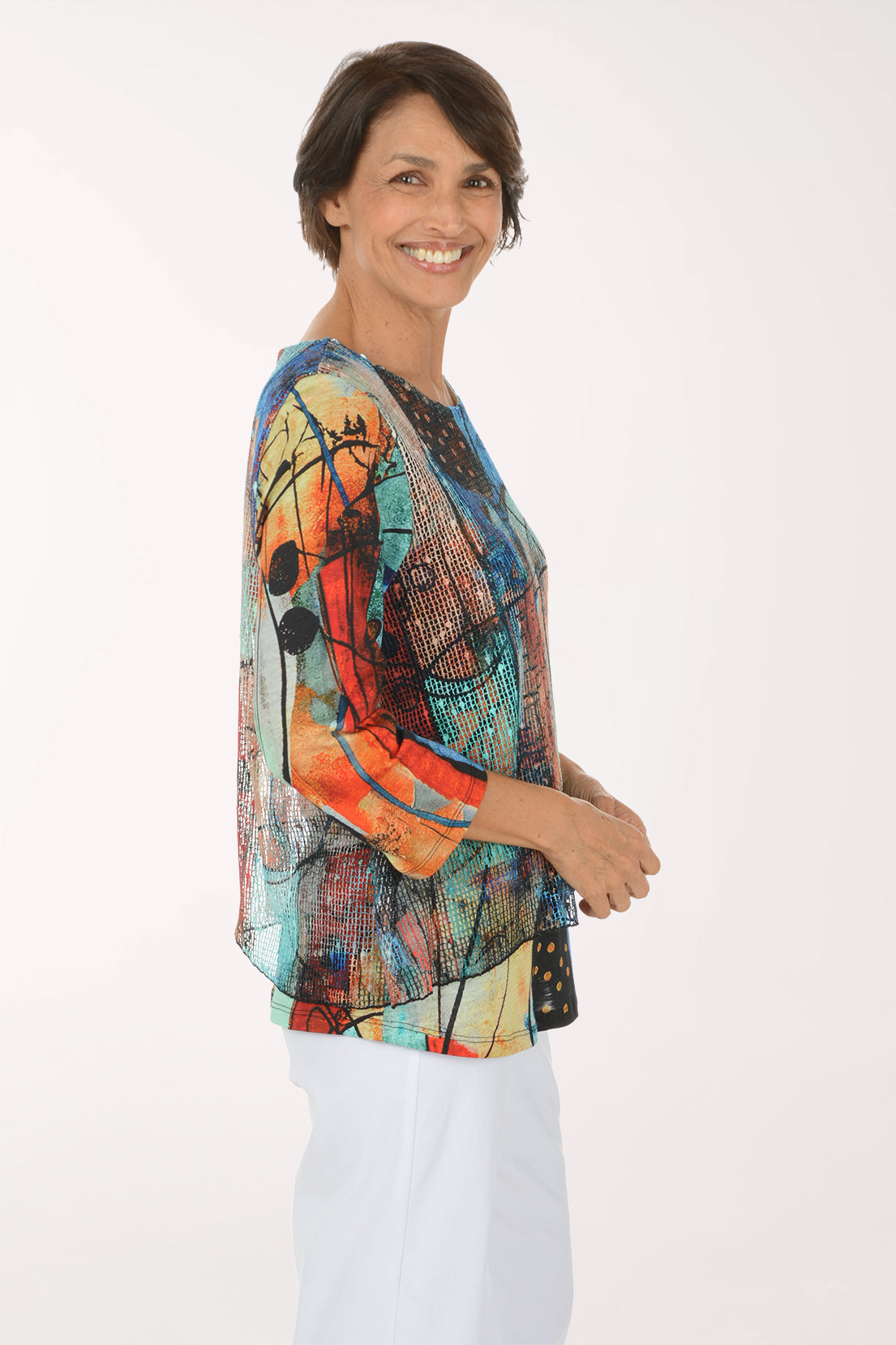 A smiling woman wearing a colorful abstract print top with three-quarter sleeves and white pants, standing against a plain white studio background.