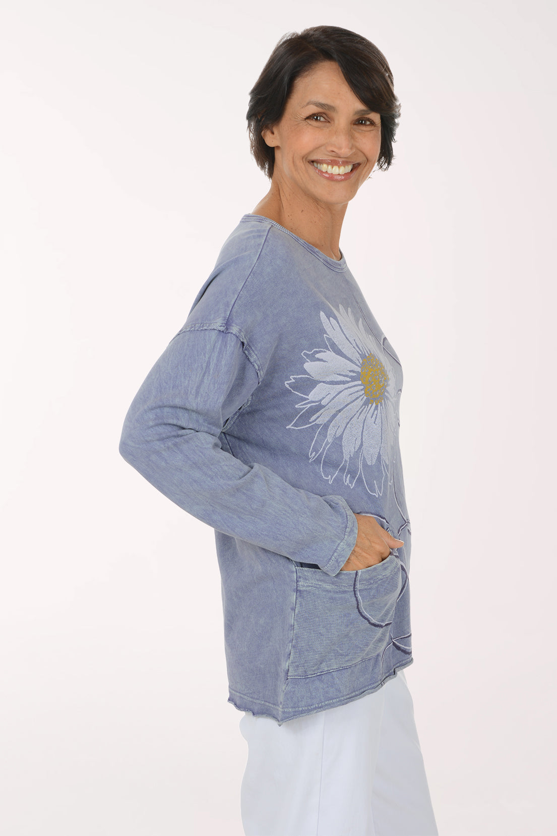 A smiling woman wearing a light blue long-sleeve top with large white daisy floral prints and white pants, standing against a plain white studio background.
