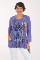 A smiling woman wearing a lavender-purple long-sleeve tunic top with a colorful abstract floral and botanical print, paired with white pants, against a plain white studio background.