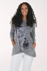 A woman with long, curly black hair wearing a gray mineral-wash long-sleeve tunic top with a purple and black floral print, paired with white pants, standing against a plain white background.