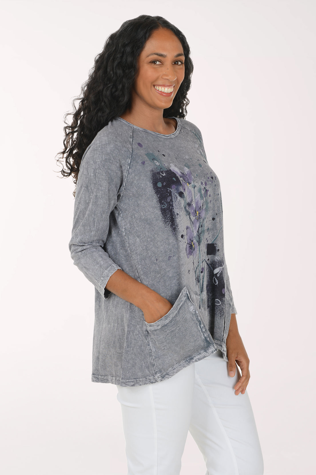 A woman with long, curly black hair wearing a gray mineral-wash long-sleeve tunic top with a purple and black floral print, paired with white pants, standing against a plain white background.