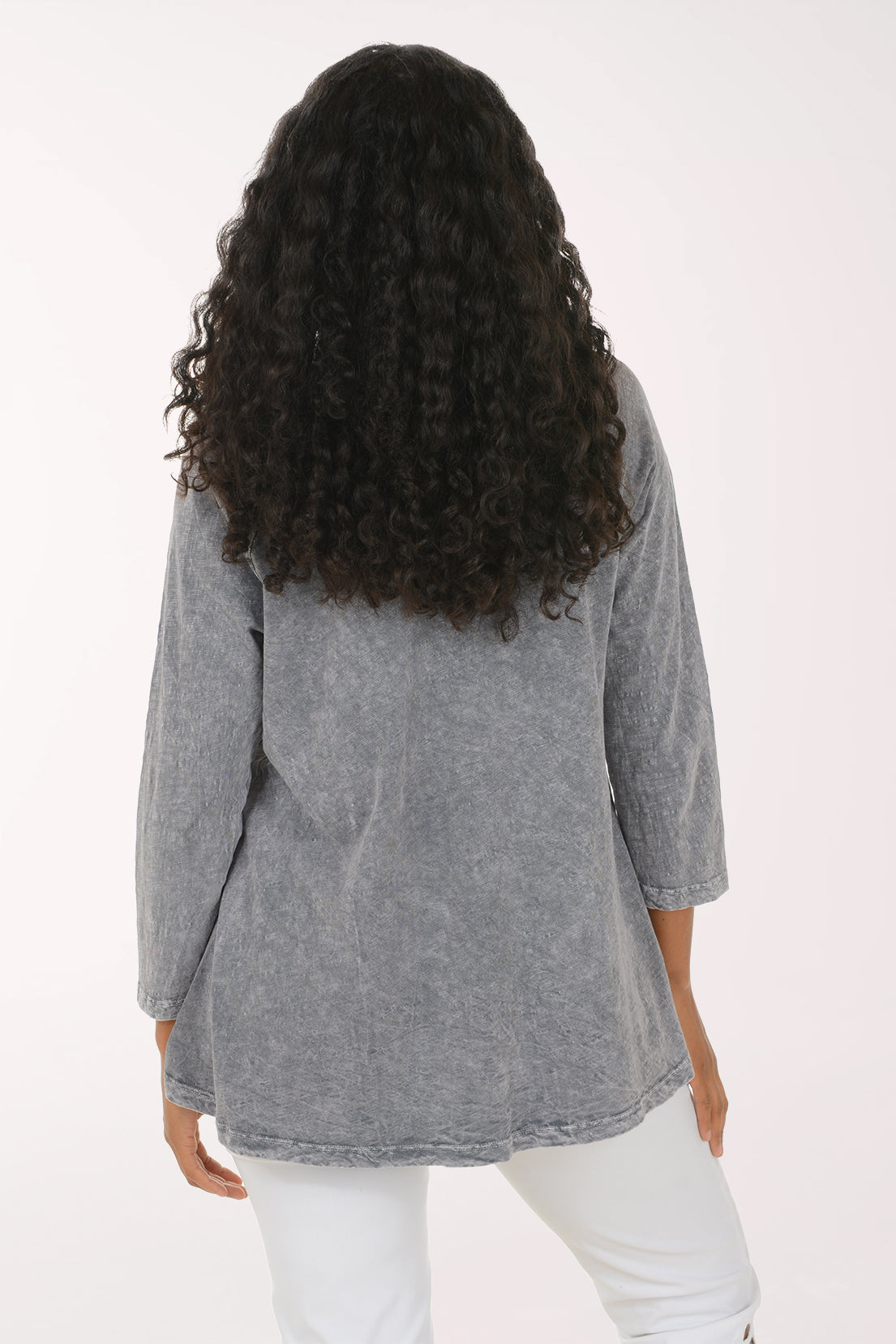 A woman with long, curly black hair wearing a gray mineral-wash long-sleeve tunic top with a purple and black floral print, paired with white pants, standing against a plain white background.