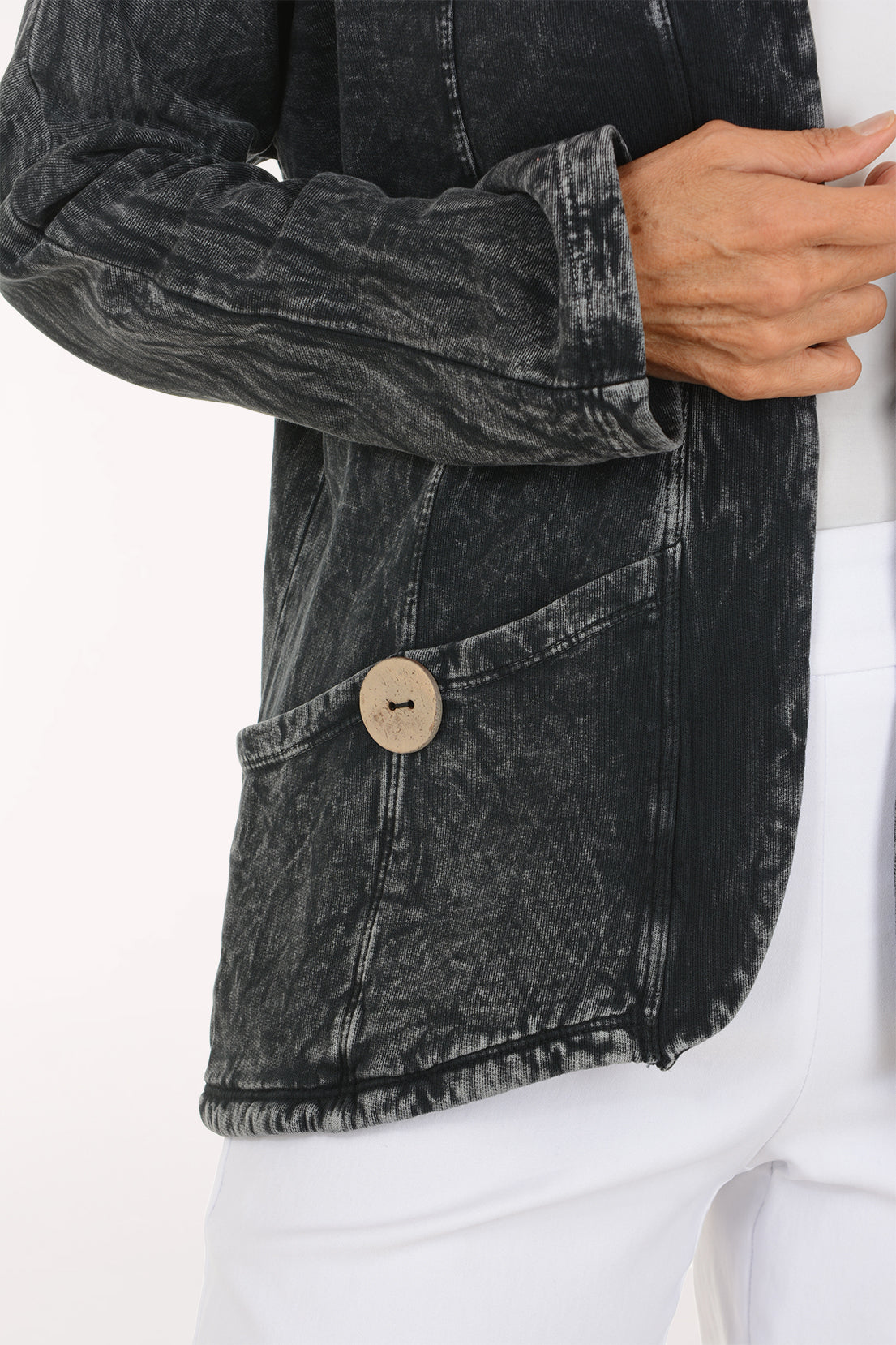 A smiling woman wearing a dark gray mineral-wash denim jacket over a white top and white pants, photographed against a clean white studio background.