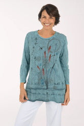 A smiling woman wearing a long-sleeve, tiered tunic top in a washed teal blue color with abstract black and red botanical line drawings, paired with white pants, against a plain white studio background.