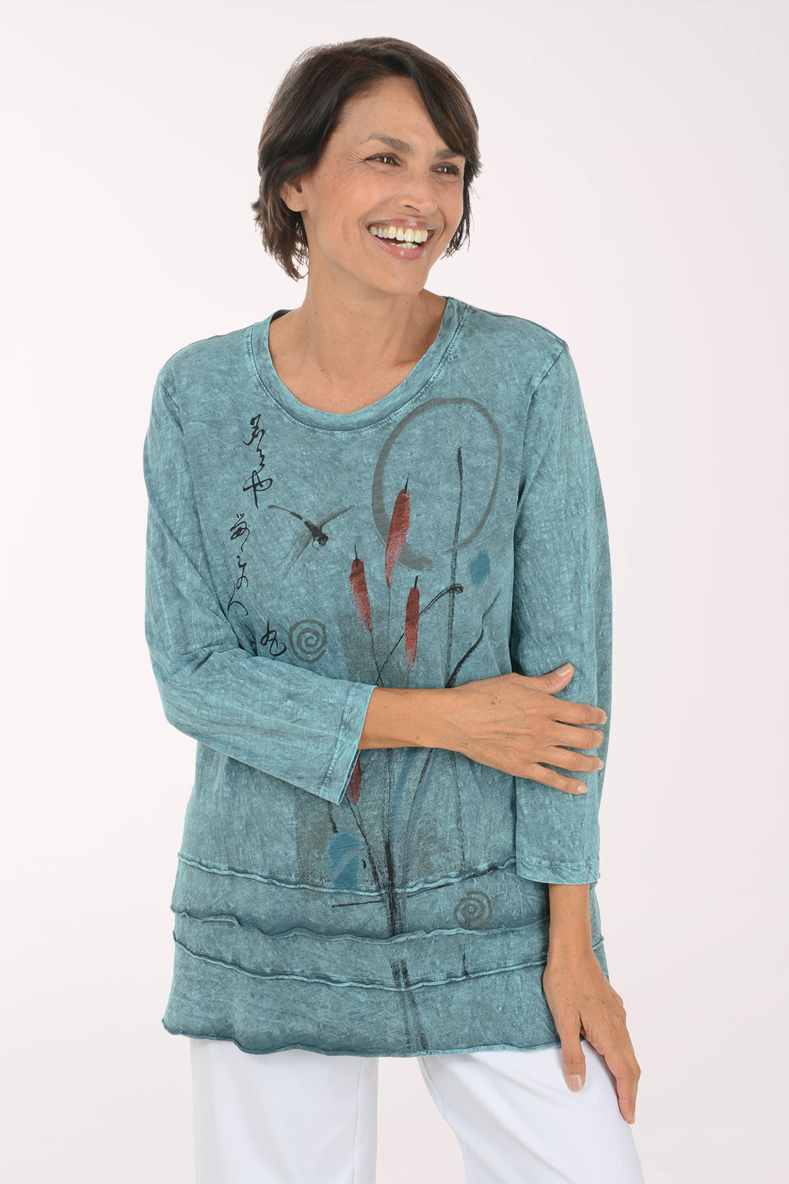 A smiling woman wearing a long-sleeve, tiered tunic top in a washed teal blue color with abstract black and red botanical line drawings, paired with white pants, against a plain white studio background.