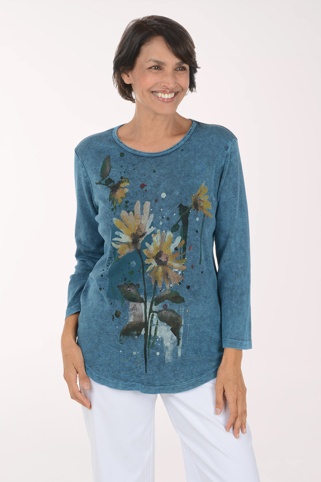 A smiling woman wearing a long-sleeve blue tunic top with a yellow and green floral print, paired with white pants, against a plain white background.