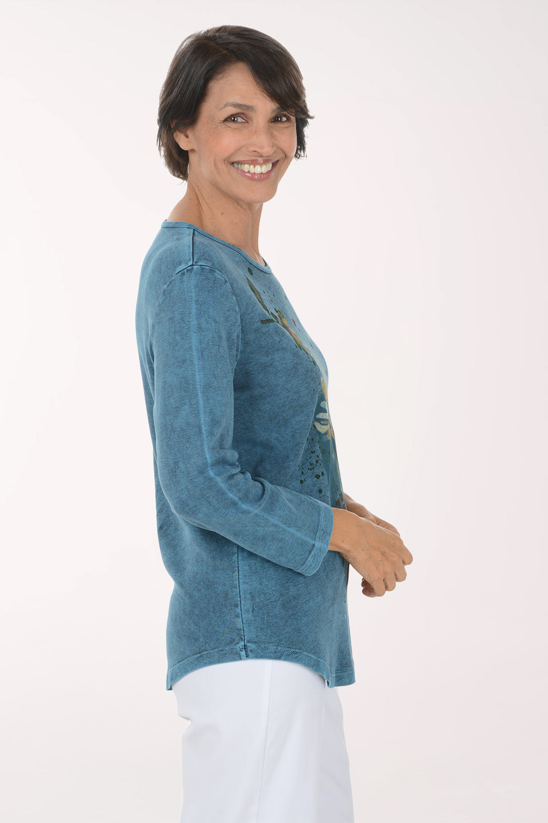 A smiling woman wearing a long-sleeve blue tunic top with a yellow and green floral print, paired with white pants, against a plain white background.