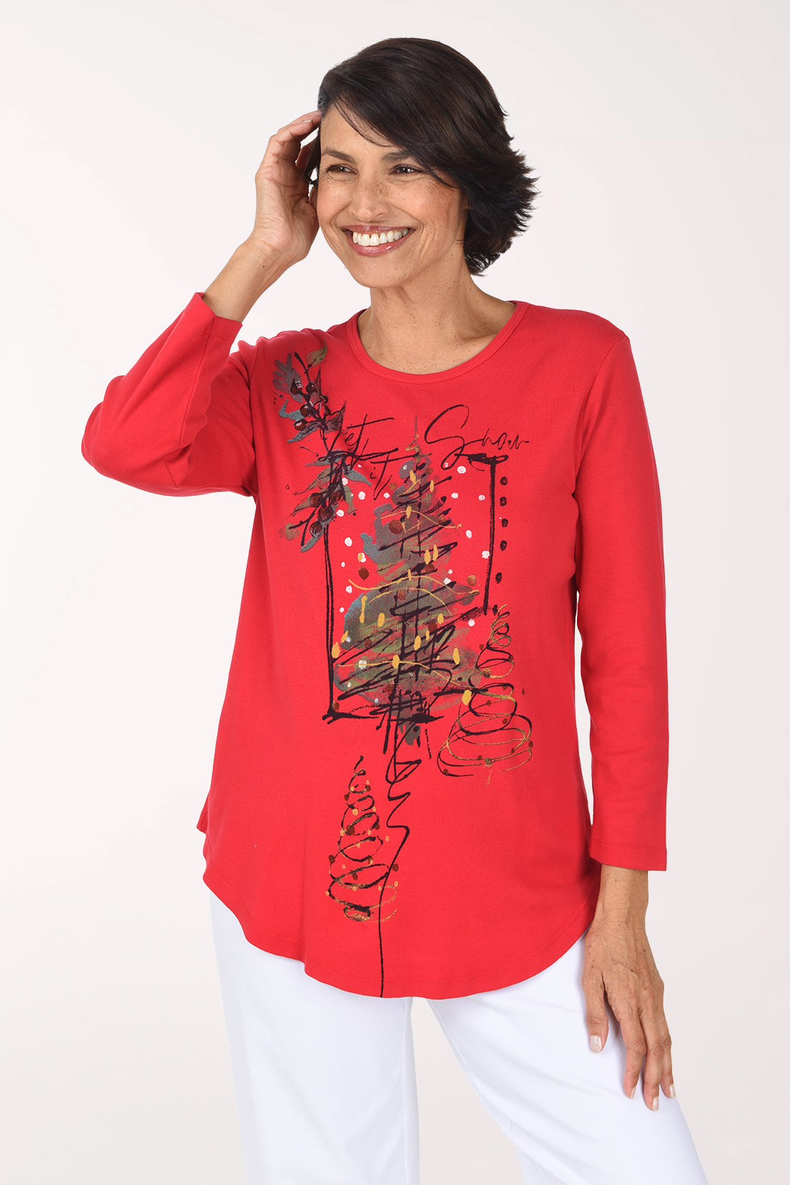 Smiling woman in a red scoop neck top with a front graphic of a sketched Christmas tree in black, gold and green with gold dots and script lettering, worn with white pants