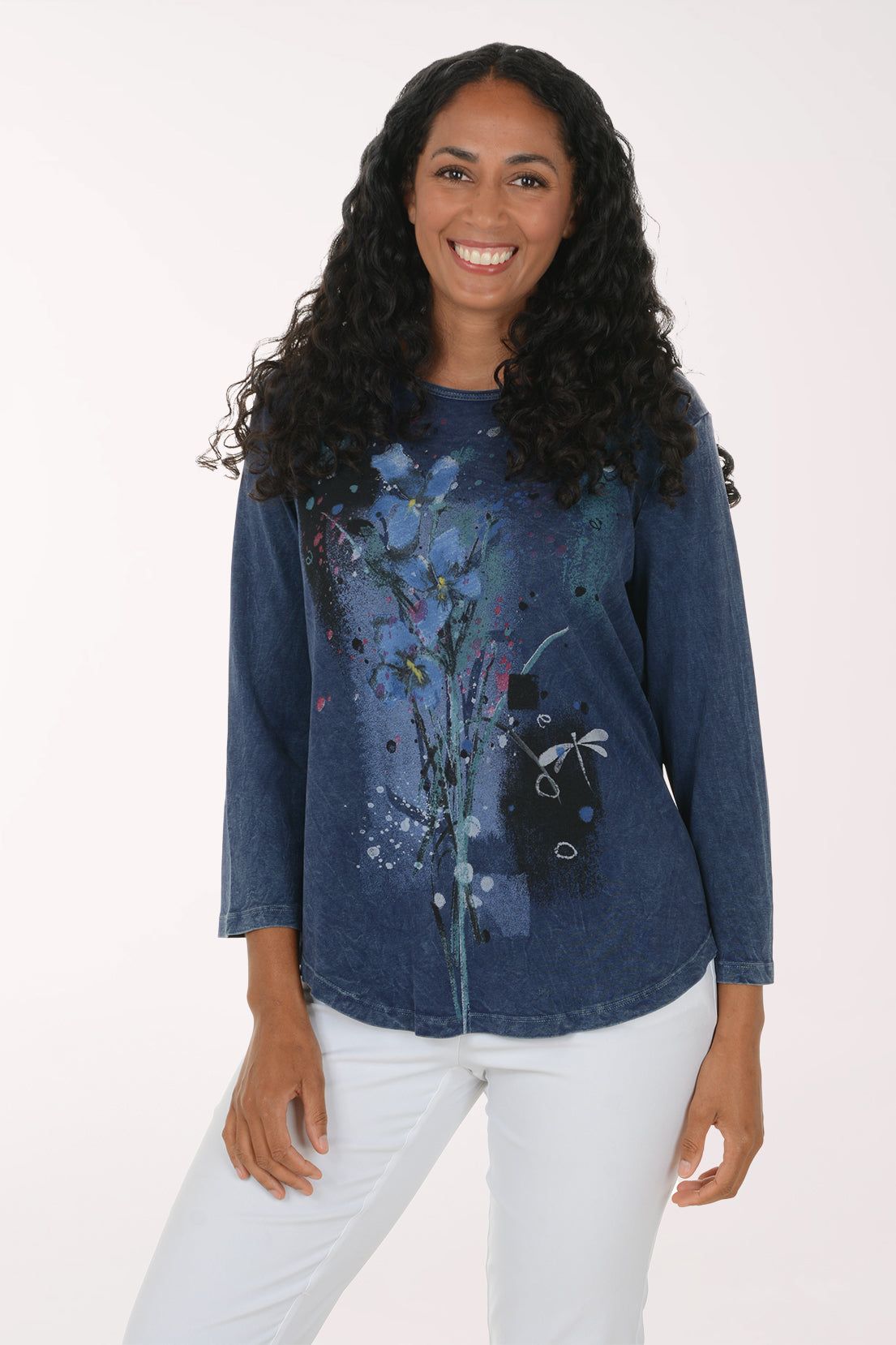 A woman with long curly black hair wearing a blue long-sleeve top with a colorful abstract floral print and white pants, standing against a plain white studio background.