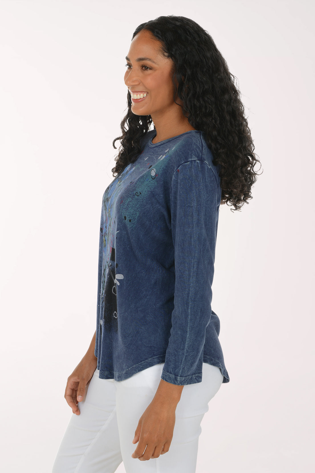 A woman with long curly black hair wearing a blue long-sleeve top with a colorful abstract floral print and white pants, standing against a plain white studio background.