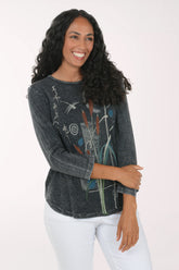 A woman with long curly black hair smiling, wearing a dark charcoal gray long-sleeve t-shirt with a colorful abstract botanical graphic print, paired with white pants, against a plain white studio background.