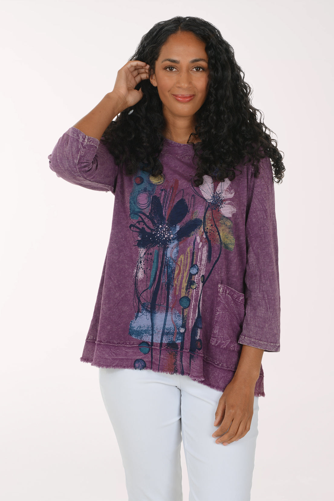 A woman modeling a purple, long-sleeved, acid-wash top with a colorful abstract floral graphic on the front, paired with white pants, against a plain white background.