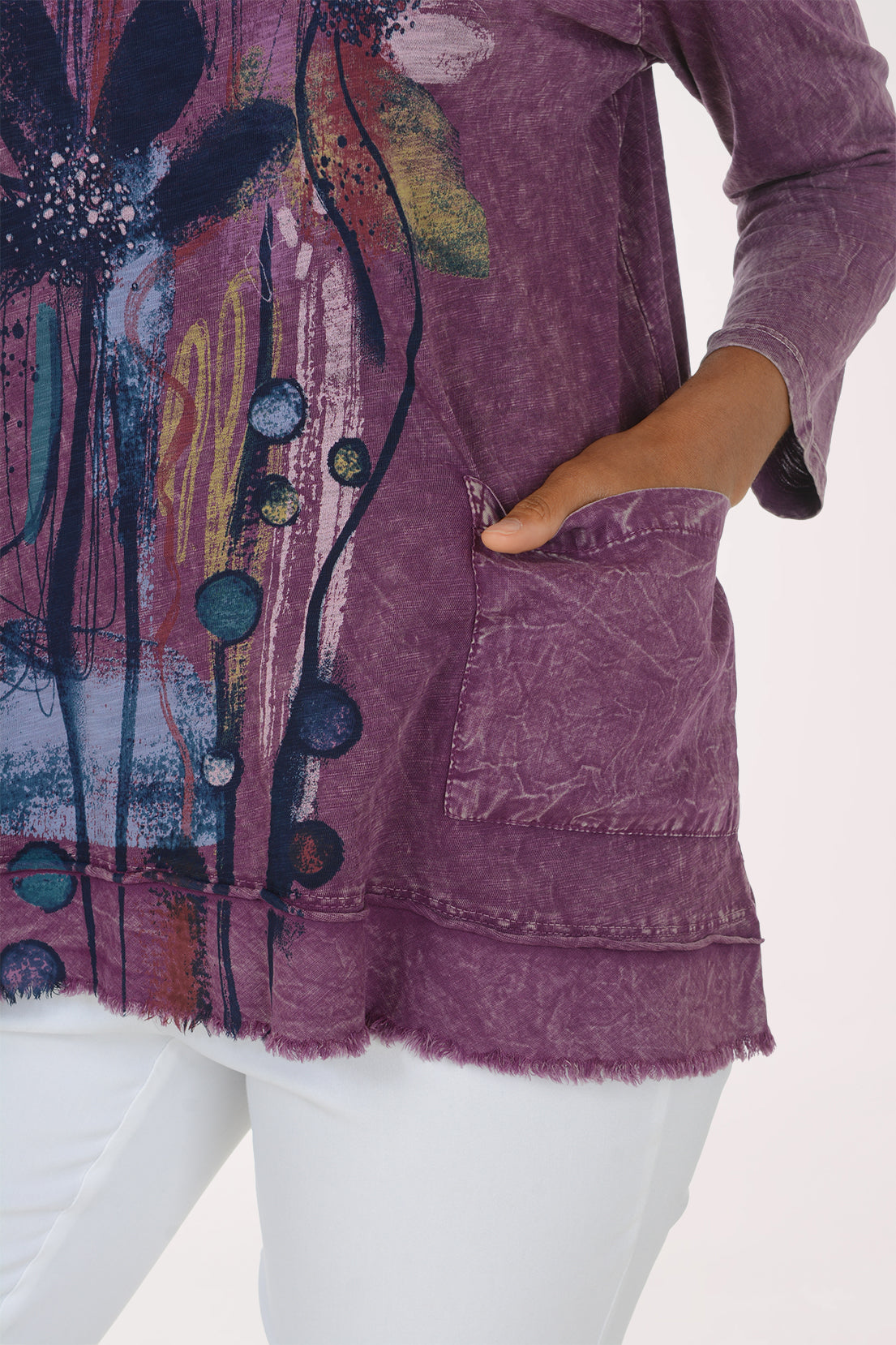 A woman modeling a purple, long-sleeved, acid-wash top with a colorful abstract floral graphic on the front, paired with white pants, against a plain white background.
