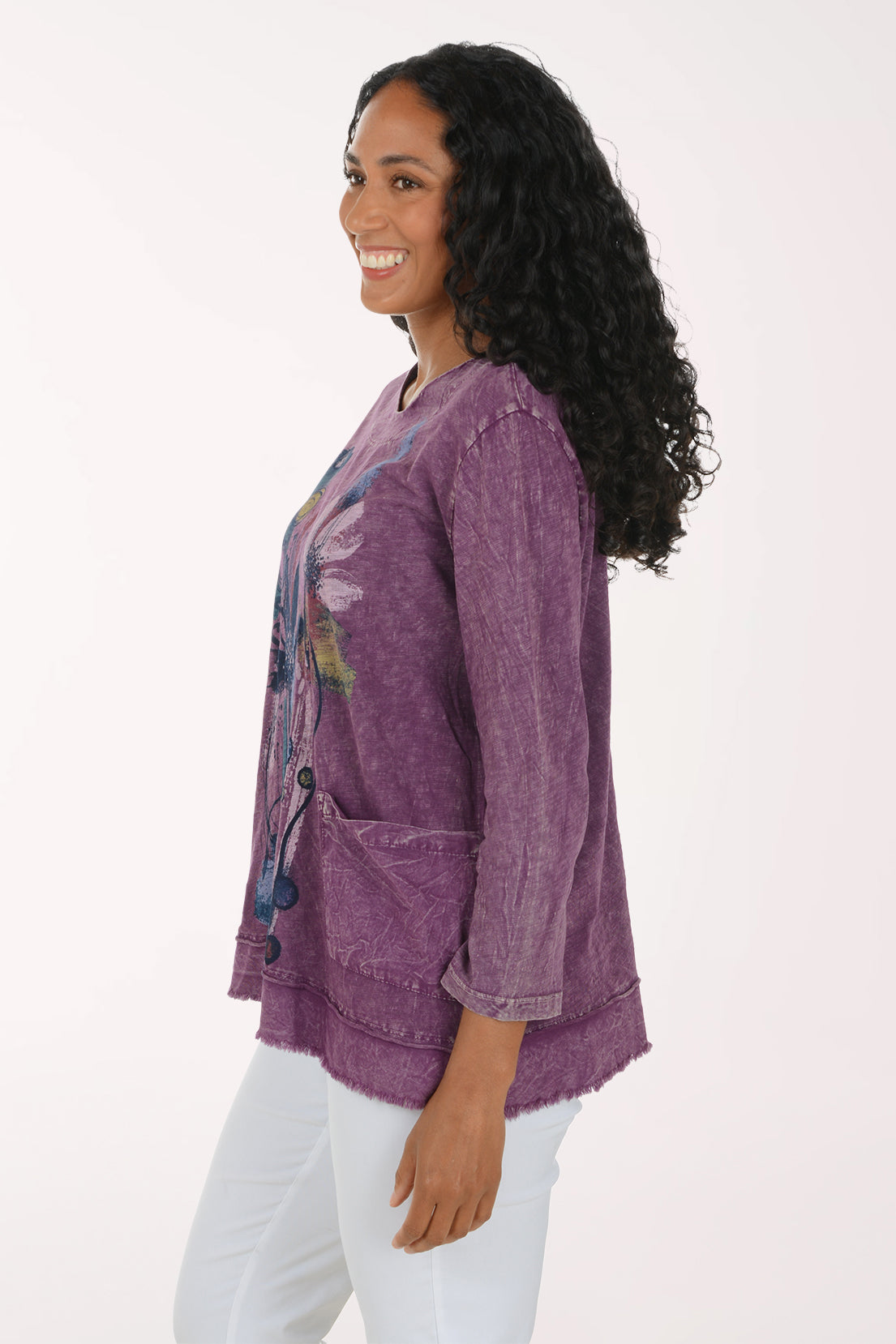 A woman modeling a purple, long-sleeved, acid-wash top with a colorful abstract floral graphic on the front, paired with white pants, against a plain white background.