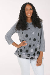 A woman with long curly black hair wearing a gray three-quarter sleeve top with a black and white polka dot pattern, paired with white pants, posing against a plain white background.