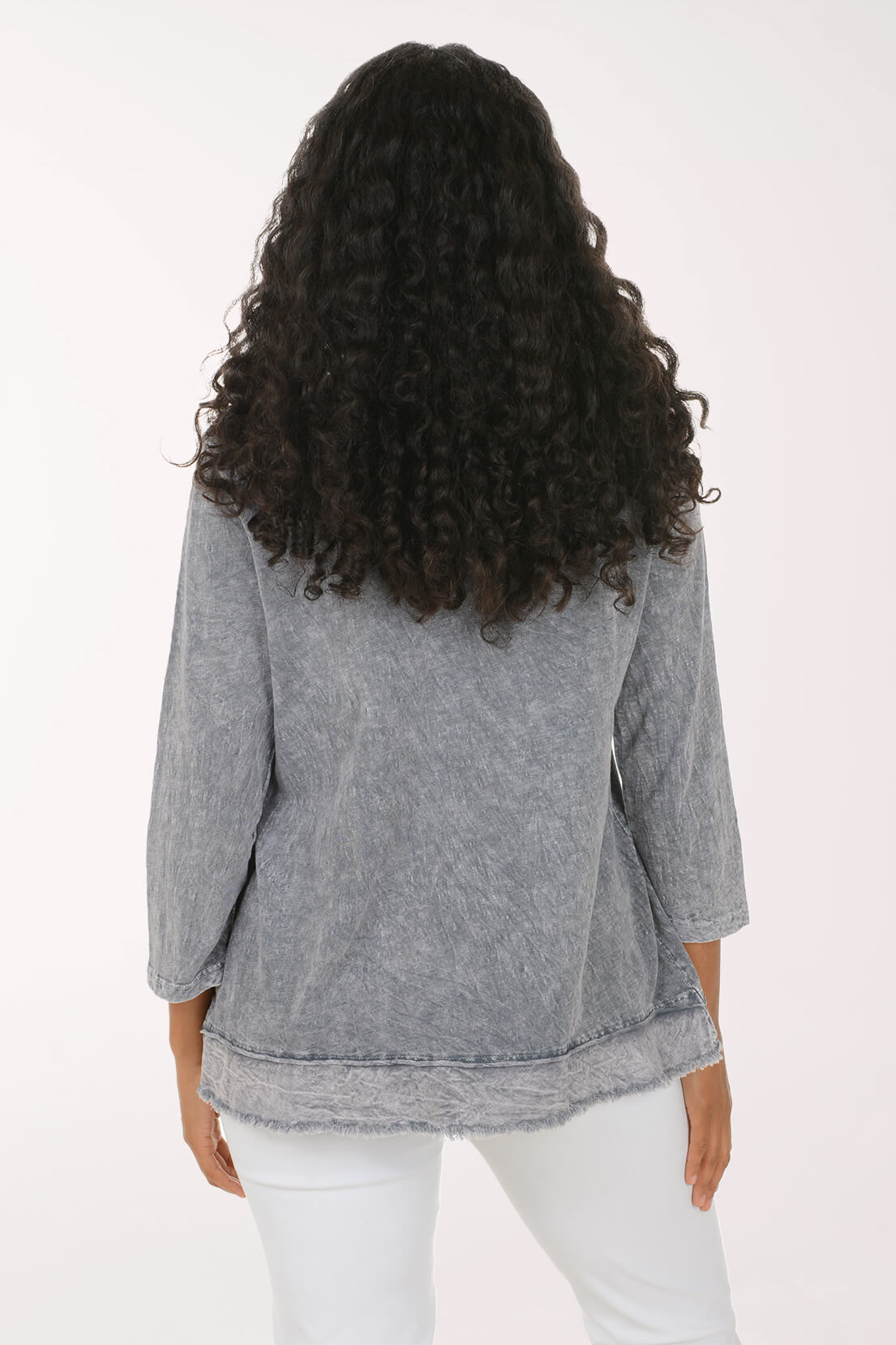 A woman viewed from the back wearing a gray three-quarter sleeve top with a raw, frayed hem and white pants, set against a plain white studio background.